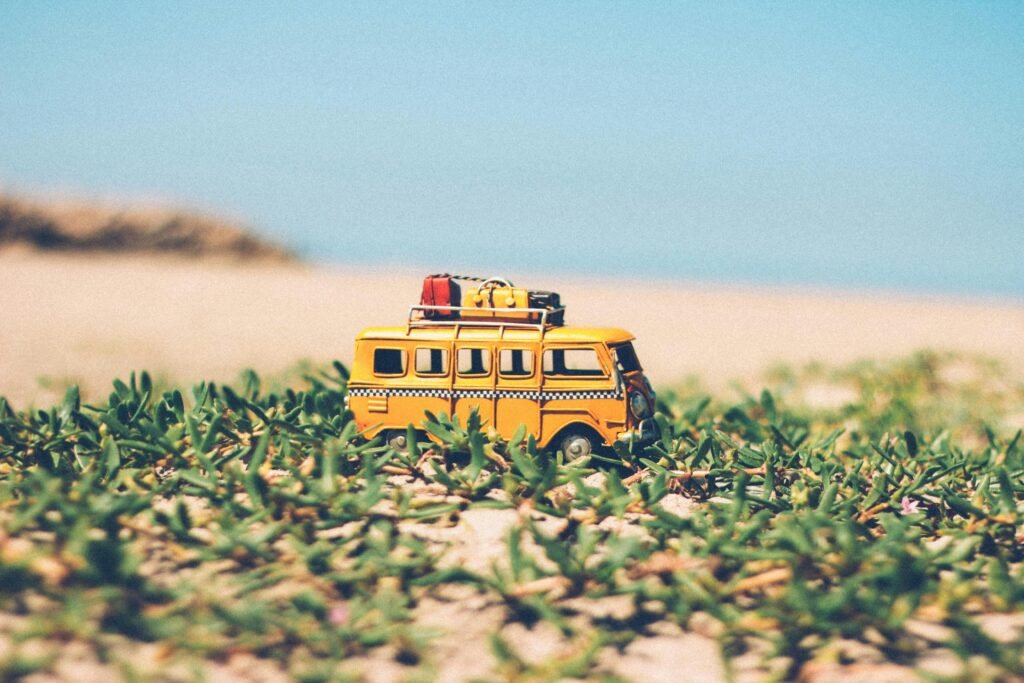 pexels-photo-386010-386010 A toy yellow camper van on beach sand surrounded by green plants under a blue sky.
